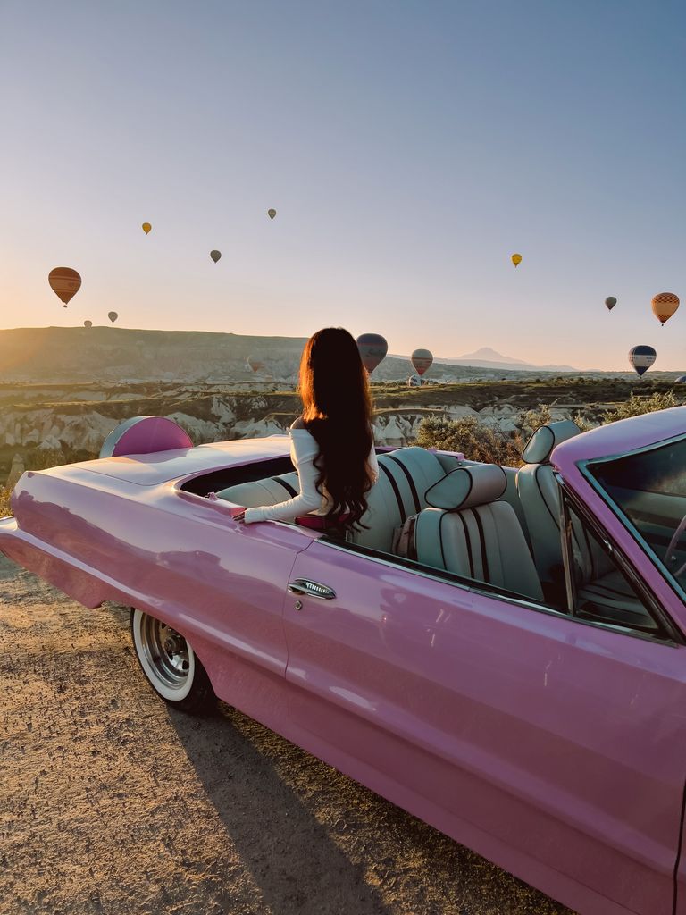 A woman in a vintage pink convertible watches hot air balloons over Cappadocia's landscape at sunset.